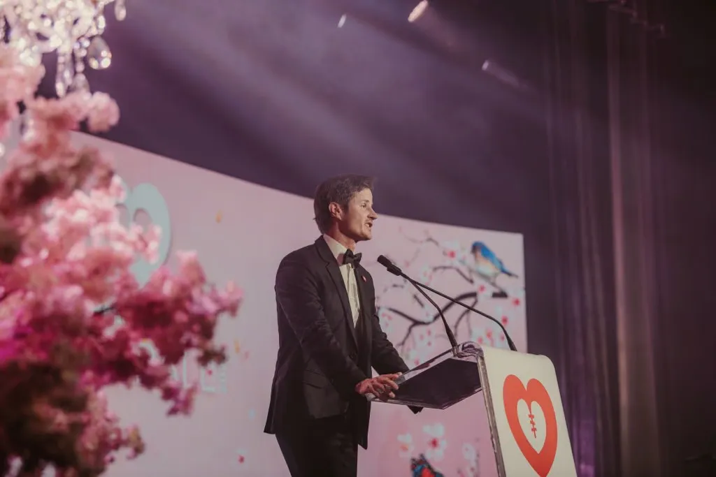 Ross Wallman presenting from a lectern on a large stage in front of a full-width screen